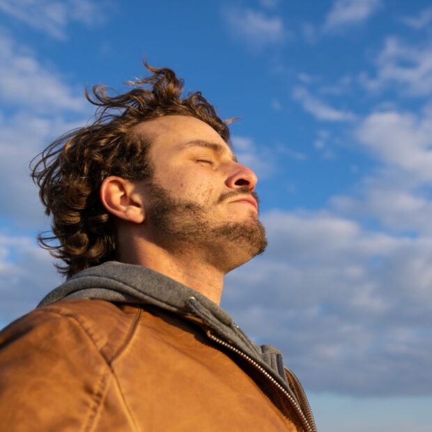 Young man outdoors, eyes closed toward warm sun and open sky, wind on his face after a hard week.