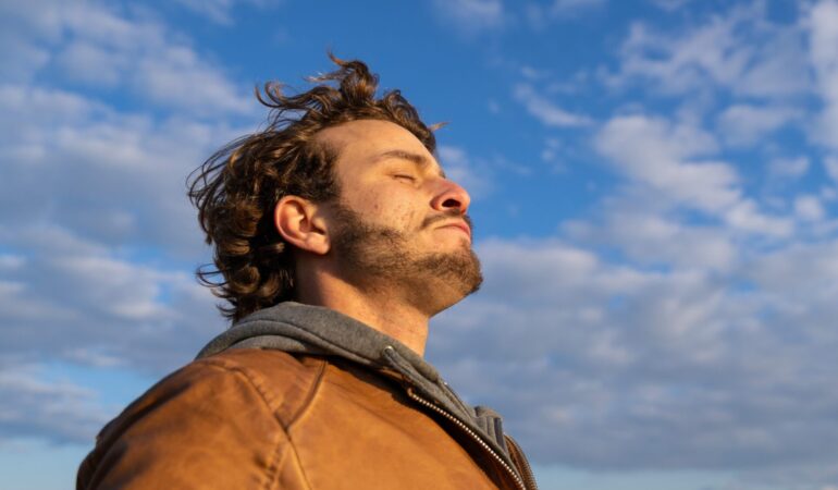 Young man outdoors, eyes closed toward warm sun and open sky, wind on his face after a hard week.