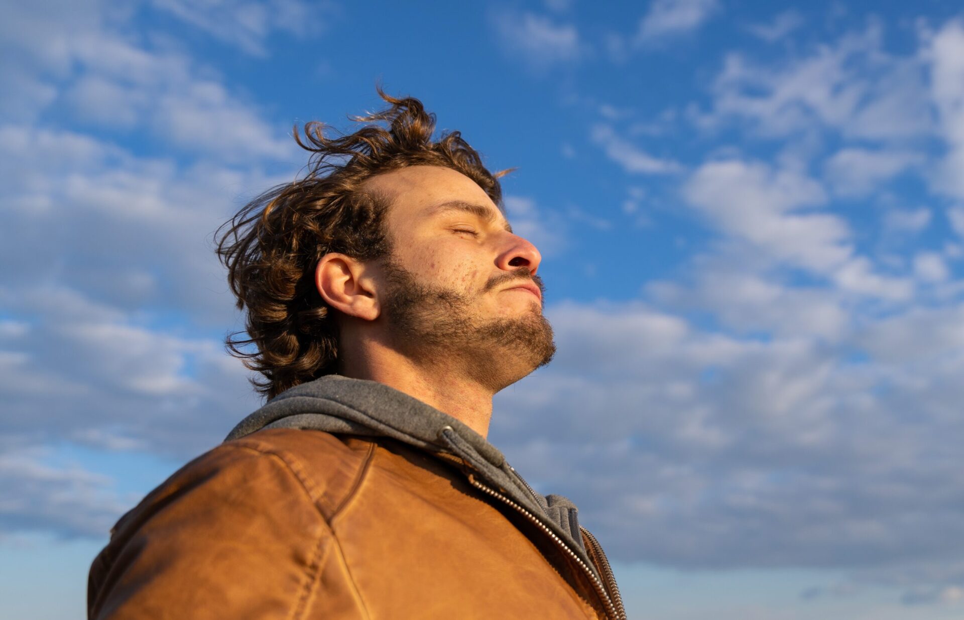 Young man outdoors, eyes closed toward warm sun and open sky, wind on his face after a hard week.
